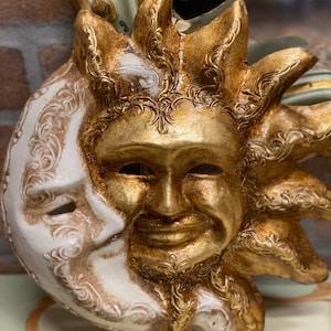 May include: A gold and white ceramic mask depicting the sun and moon. The sun is smiling and has a golden face with intricate details. The moon is white and has a more serious expression.