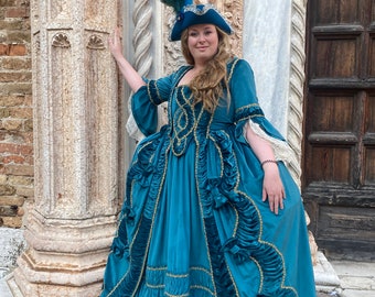 May include: A woman wearing a teal blue velvet gown with gold trim and a blue feathered hat. The gown has a ruffled skirt and a fitted bodice. She is standing in front of a stone wall.