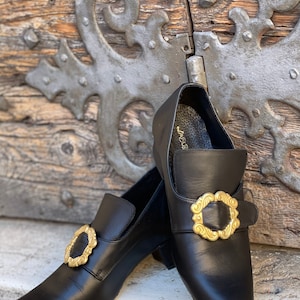 May include: A pair of black leather shoes with gold buckles. The shoes have a square toe and a low heel. The buckles are ornate and have a floral design.
