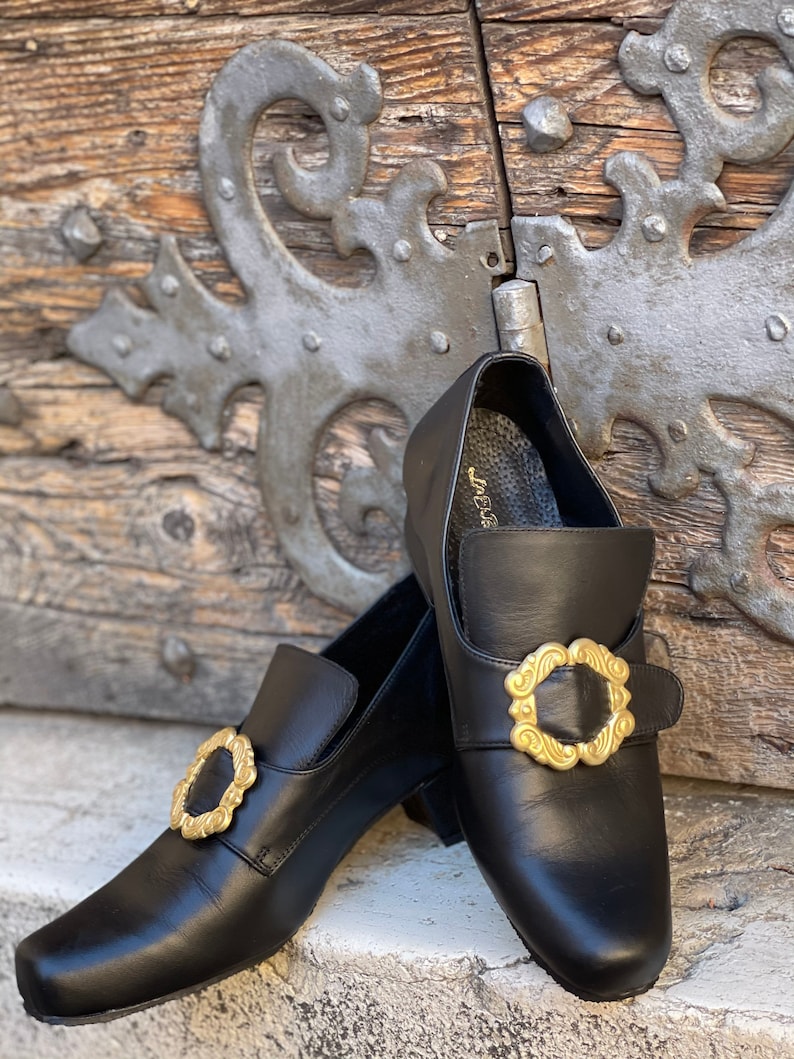 May include: A pair of black leather shoes with gold buckles. The shoes have a square toe and a low heel. The buckles are ornate and have a floral design.