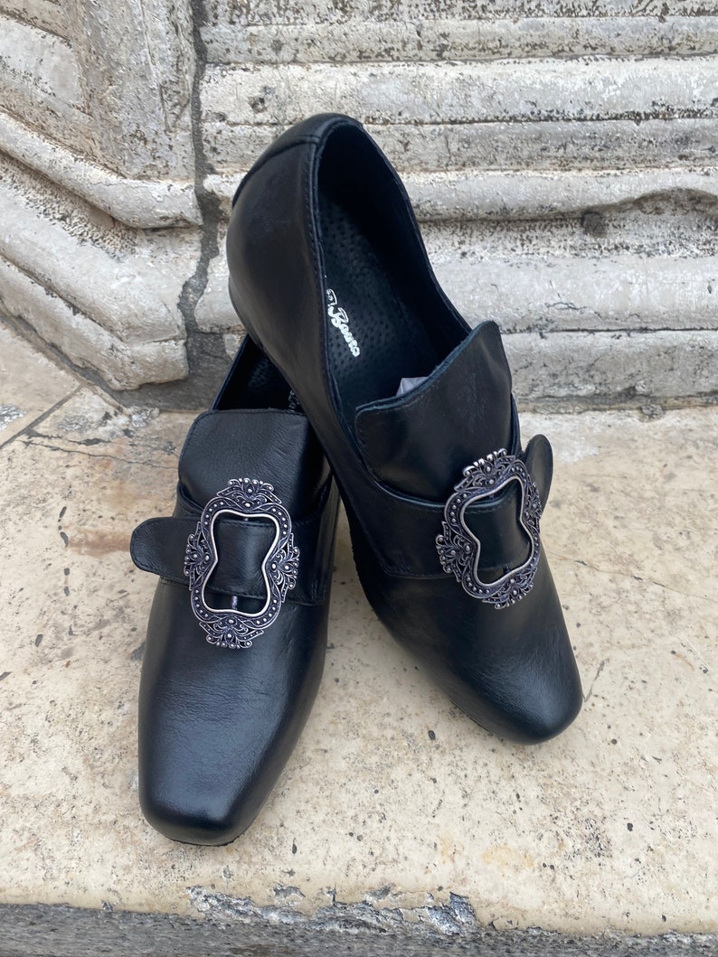 May include: Black leather shoes with silver buckles. The shoes have a square toe and a low heel.