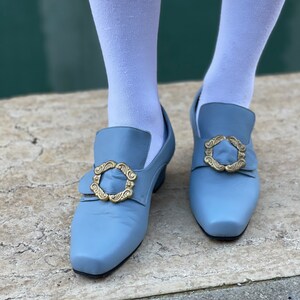 Puede incluir: Un par de zapatos de cuero azul claro con hebillas doradas. Los zapatos tienen una punta puntiaguda y un tac&oacute;n bajo.