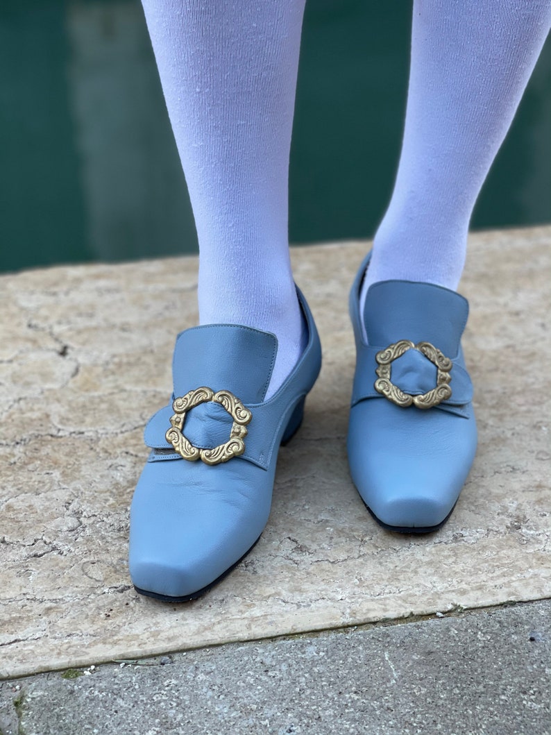 Puede incluir: Un par de zapatos de cuero azul claro con hebillas doradas. Los zapatos tienen una punta puntiaguda y un tac&oacute;n bajo.