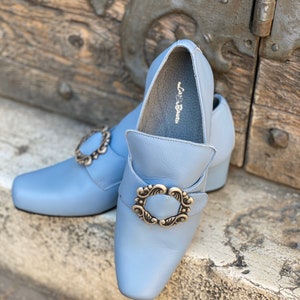 Puede incluir: Un par de zapatos de cuero azul claro con una hebilla plateada decorativa. Los zapatos tienen una punta puntiaguda y un tac&oacute;n bajo. El nombre de la marca "La Botte" es visible en el interior de un zapato.