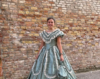 May include: A woman in a sage green gown with white lace trim and a matching handbag stands in front of a brick wall. She is wearing a tiara and a pearl necklace.