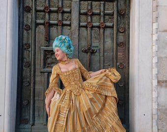 May include: A woman wearing a blue wig and a gold and white striped dress stands in front of a wooden door with a date carved above it. The door is decorated with metal flowers.