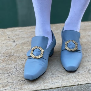 Puede incluir: Un par de zapatos azul claro con hebillas doradas. Los zapatos tienen un tac&oacute;n bajo y una punta puntiaguda.
