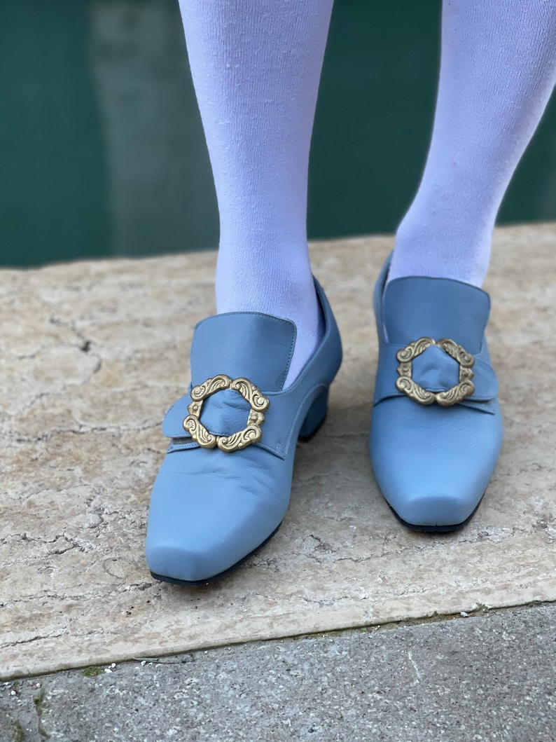 Puede incluir: Un par de zapatos azul claro con hebillas doradas. Los zapatos tienen un tac&oacute;n bajo y una punta puntiaguda.