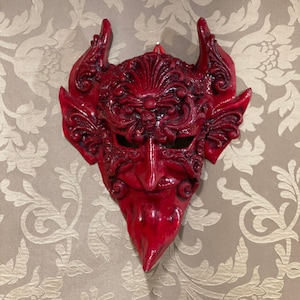 May include: A red devil mask with intricate details and horns. The mask is made of a hard material and has a textured surface.