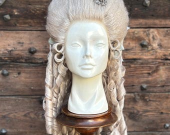 May include: A vintage-style wig on a mannequin head. The wig is light gray with large, curled ringlets and a high, bouffant style. A decorative silver and turquoise brooch adorns the top. The mannequin is displayed on a dark wooden stand.