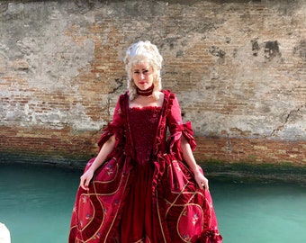 Könnte beinhalten: Ein bodenlanges, dunkelrotes Kleid mit strukturiertem Oberteil, Rüschenbesatz und goldfarbenen Paspeln. Der Rock hat eine voluminöse Silhouette mit Blumenstickereien, ergänzt durch ein passendes Halsband und einen kleinen Beutel mit Kordelzug.