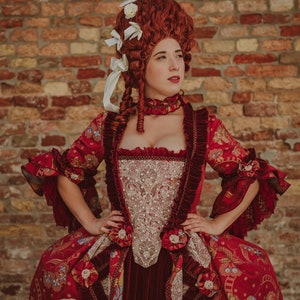 May include: A woman in a red and gold brocade dress with a white lace bodice and a white lace trim. She is wearing a red wig with white flowers and a red choker necklace. She is standing in front of a brick wall.