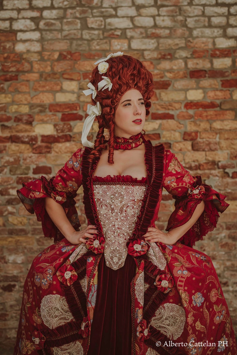 May include: A woman in a red and gold brocade dress with a white lace bodice and a white lace trim. She is wearing a red wig with white flowers and a red choker necklace. She is standing in front of a brick wall.