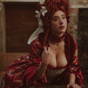 May include: A woman in a red and gold brocade dress with a white lace collar and a white and red wig with white flowers. She is leaning on a large, rusty metal chest.