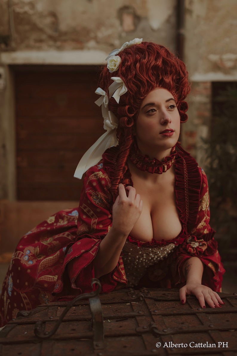 May include: A woman in a red and gold brocade dress with a white lace collar and a white and red wig with white flowers. She is leaning on a large, rusty metal chest.
