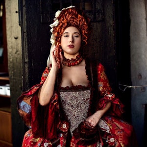 May include: A woman in a red and gold brocade dress with a white lace bodice and a large, curly red wig. She is wearing a white and gold necklace and has a serious expression on her face.