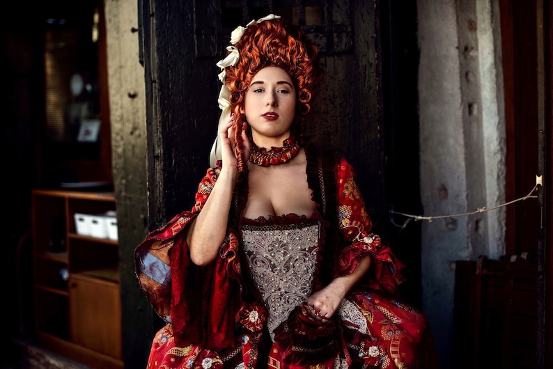 May include: A woman in a red and gold brocade dress with a white lace bodice and a large, curly red wig. She is wearing a white and gold necklace and has a serious expression on her face.