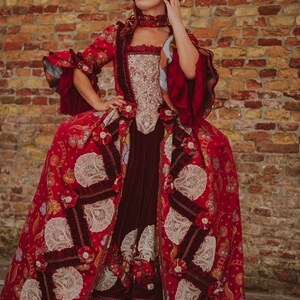 May include: A woman in a red and white gown with lace details and a red wig. The gown has a fitted bodice and a full skirt. The woman is standing in front of a brick wall.