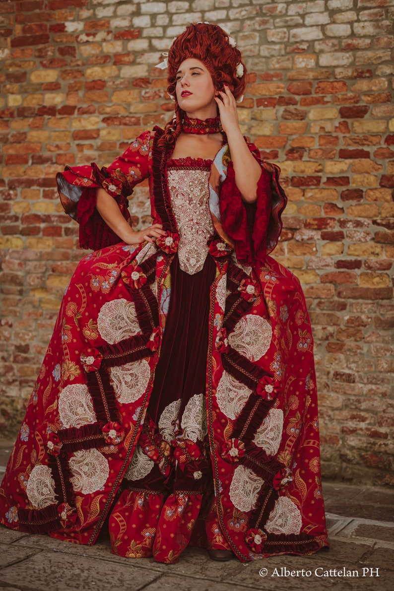 May include: A woman in a red and white gown with lace details and a red wig. The gown has a fitted bodice and a full skirt. The woman is standing in front of a brick wall.