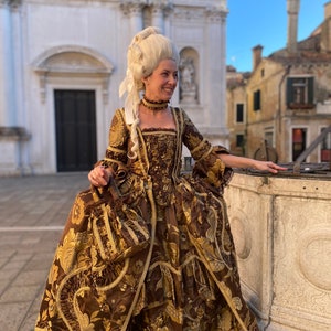 May include: A woman wearing a brown and gold brocade gown with a white lace trim. She is wearing a white wig and a white lace bow around her neck. She is standing in front of a building with a white facade.