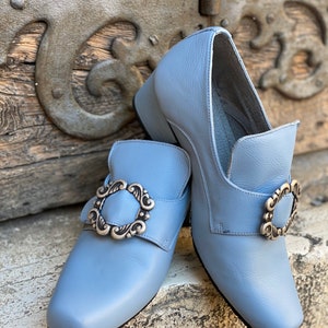 Puede incluir: Un par de zapatos de cuero azul claro con hebillas plateadas. Los zapatos tienen un tac&oacute;n bajo y una punta puntiaguda.
