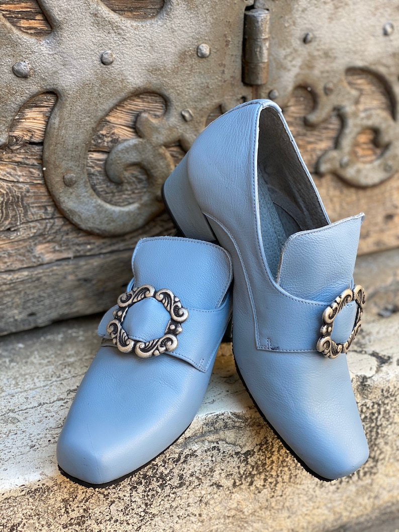 Puede incluir: Un par de zapatos de cuero azul claro con hebillas plateadas. Los zapatos tienen un tac&oacute;n bajo y una punta puntiaguda.