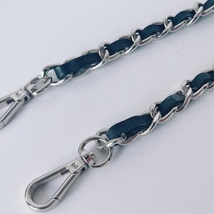Könnte beinhalten: Zwei silberfarbene Metallkettenriemen mit schwarzen Lederakzenten. Jeder Riemen hat ein geflochtenes Kettendesign und einen silbernen Verschluss an jedem Ende. Die Riemen sind wahrscheinlich für eine Handtasche.
