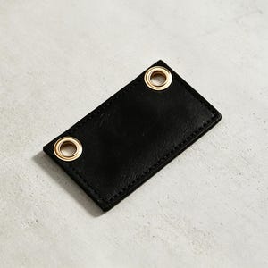 以下が含まれることがあります： 対角線上の角に2つの金色の金属製ハトメが付いた、縁が縫製された長方形の黒いタグ。タグは滑らかで平らな表面と、すっきりとした均一な縁取りが特徴で、取り付けやラベル付けに適した機能的なデザインとなっています。