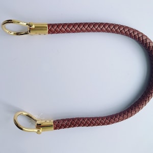 Puede incluir: Un asa de bolso de cuero trenzado marrón con cierres metálicos dorados. El asa tiene forma de U, con una textura tejida. Los cierres dorados están unidos a cada extremo del asa.