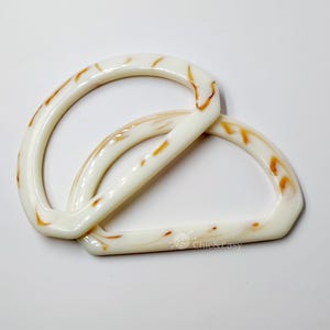 May include: Two white plastic D-shaped handles with a marbled pattern in shades of brown and white. The handles are stacked on top of each other.