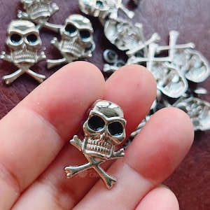 May include: Silver skull charm with black eye sockets and crossed bones. The charm has a detailed skull design, possibly for jewellery or crafts. Other similar charms are visible in the background.