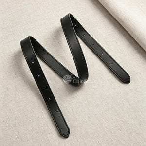 Könnte beinhalten: Ein schmaler schwarzer Gürtel mit glatter Oberfläche und einer Reihe gleichmäßig verteilter Verstelllöcher entlang des Riemens. Der Gürtel ist in einer geschwungenen, überlappenden Form angeordnet und zeigt das flexible Material und das spitze Ende.