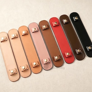 May include: Eight rectangular leather handle covers arranged in a row, featuring a gradient of colors from light beige to black. Each cover has two small loops secured with gold-toned metal snap buttons at the top and bottom ends.