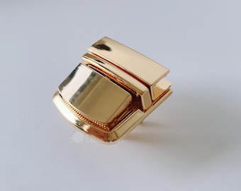May include: A shiny, gold-colored metal clasp with a rectangular shape and a decorative, textured edge. The clasp has a geometric design with multiple layers and a polished finish, suitable for bags or accessories.