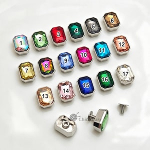 Puede incluir: Una colección de diecisiete botones rectangulares de cristal facetado en marcos de metal plateado, numerados del 1 al 17. Los botones presentan varios colores, incluyendo iridiscente, verde, rojo, negro, dorado, azul, rosa y transparente, con tornillos.