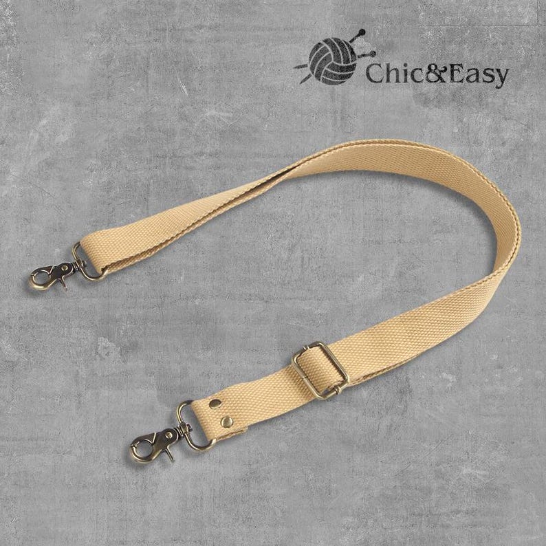 May include: A beige canvas adjustable strap with two metal clasps and a metal slider. The strap is for a bag or purse.