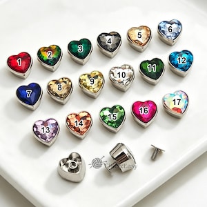May include: A collection of heart-shaped metal buttons with faceted crystals in various colors, numbered 1 through 17. Each button features a silver-tone metal base and a screw-back attachment mechanism, displayed on a white surface.