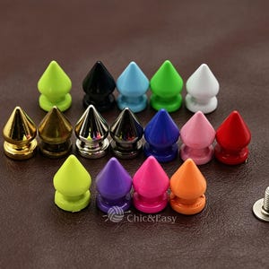 Può includere: Una collezione di borchie metalliche a forma di cono in vari colori, tra cui verde lime, nero, azzurro, bianco, oro, argento, grigio scuro, blu, rosa, rosso, viola, magenta e arancione, con una piccola vite di fissaggio visibile in basso a destra.
