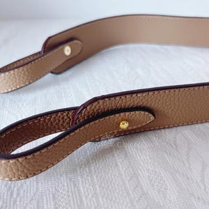 以下が含まれることがあります： テクスチャード加工が施されたタンレザーのハンドバッグストラップ。ストラップは濃い茶色のエッジが特徴で、ゴールドカラーの金属製ハードウェアで固定されています。ステッチは薄いベージュ色です。