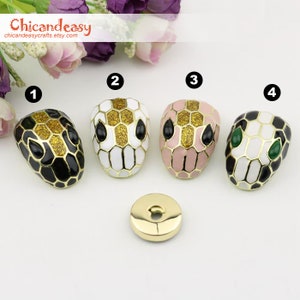 May include: Four snake head-shaped decorative buttons in black, white, and pink with gold accents. The buttons have black or green eyes and are arranged with a small gold button. The Etsy shop name "Chicandeasycrafts" is in the top left corner.