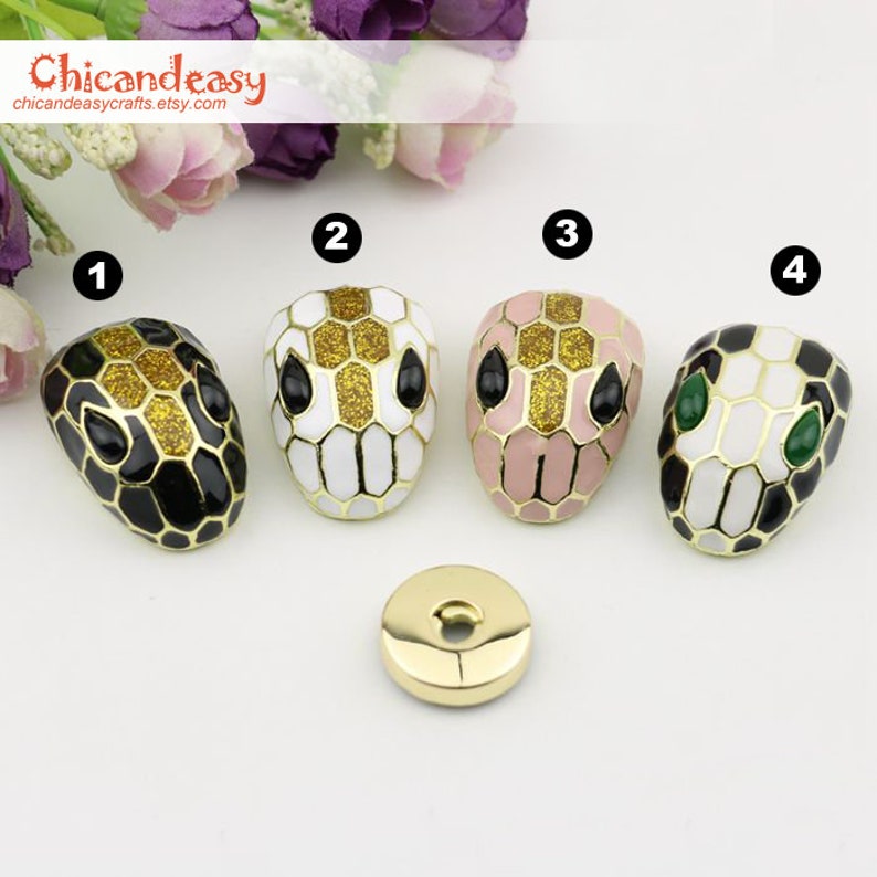 May include: Four snake head-shaped decorative buttons in black, white, and pink with gold accents. The buttons have black or green eyes and are arranged with a small gold button. The Etsy shop name "Chicandeasycrafts" is in the top left corner.