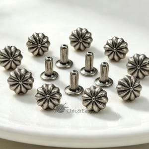 May include: A collection of silver-toned metal rivets featuring a circular, fluted, flower-like design on the head and a smooth, cylindrical post. The text Chic&Easy is visible at the bottom centre of the arrangement of these hardware fasteners.