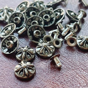 Könnte beinhalten: Eine Ansammlung von silberfarbenen Metallnieten mit einem erhabenen, mehrblättrigen Blumen- oder sternförmigen Design auf der Kappe. Jede Niete besteht aus einem dekorativen Oberteil und einem separaten Metallstift, die lose verstreut sind.