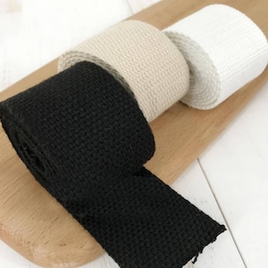 Puede incluir: Tres rollos de cinta de algod&oacute;n tejido en negro, beige y blanco. La cinta es una tira plana y estrecha de tela con una superficie texturizada.
