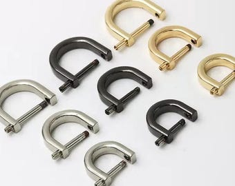 May include: A collection of D-ring hardware in various metallic finishes, including gold, silver, and black. The D-rings are arranged on a white surface, showcasing their shape and design. These are commonly used in bag making and leathercraft.