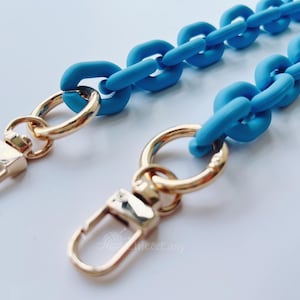 May include: A close-up shot of a light blue chain strap with gold-colored clasps and rings. The chain links are large and rounded. The clasps are rectangular with a curved opening.