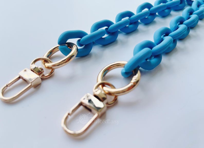May include: A close-up shot of a light blue chain strap with gold-colored clasps and rings. The chain links are large and rounded. The clasps are rectangular with a curved opening.