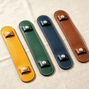Puede incluir: Cuatro fundas de cuero alargadas para asas en amarillo, verde, azul marino y marrón. Cada funda presenta costuras blancas en contraste a lo largo de los bordes y dos botones de presión metálicos plateados. El texto Chic&Easy es visible.