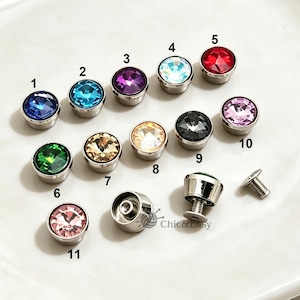 Può includere: Una collezione di undici bottoni rotondi in metallo con centri in cristallo sfaccettato multicolore. Ogni bottone è numerato da 1 a 11 e presenta varie tonalità tra cui blu, viola, rosso, verde, rosa e trasparente. I bottoni hanno una chiusura a vite.