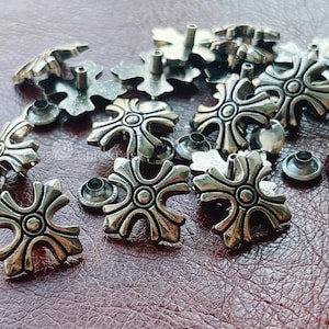 May include: A collection of silver-toned metal rivets featuring a four-pointed cross design with a central circular detail. Each rivet has a small protruding post on the reverse side, designed for attachment to fabric or leather materials.